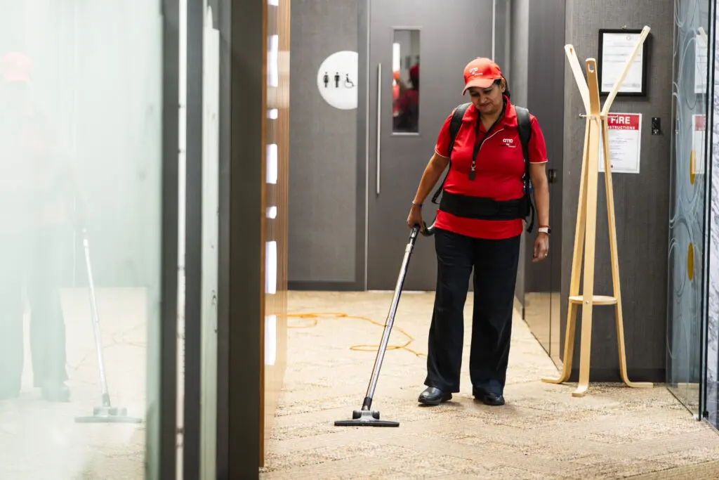 Professional cleaner in AMC Commercial Cleaning uniform vacuuming the floor demonstrating office cleaning and hygiene maintenance in a modern workspace.