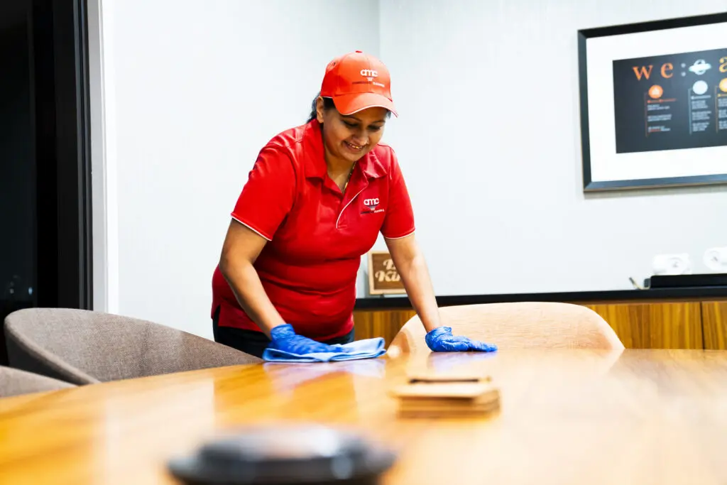 Professional cleaner in AMC Commercial Cleaning uniform wiping down a conference room table with a cloth, demonstrating office cleaning and hygiene maintenance in a modern workspace.