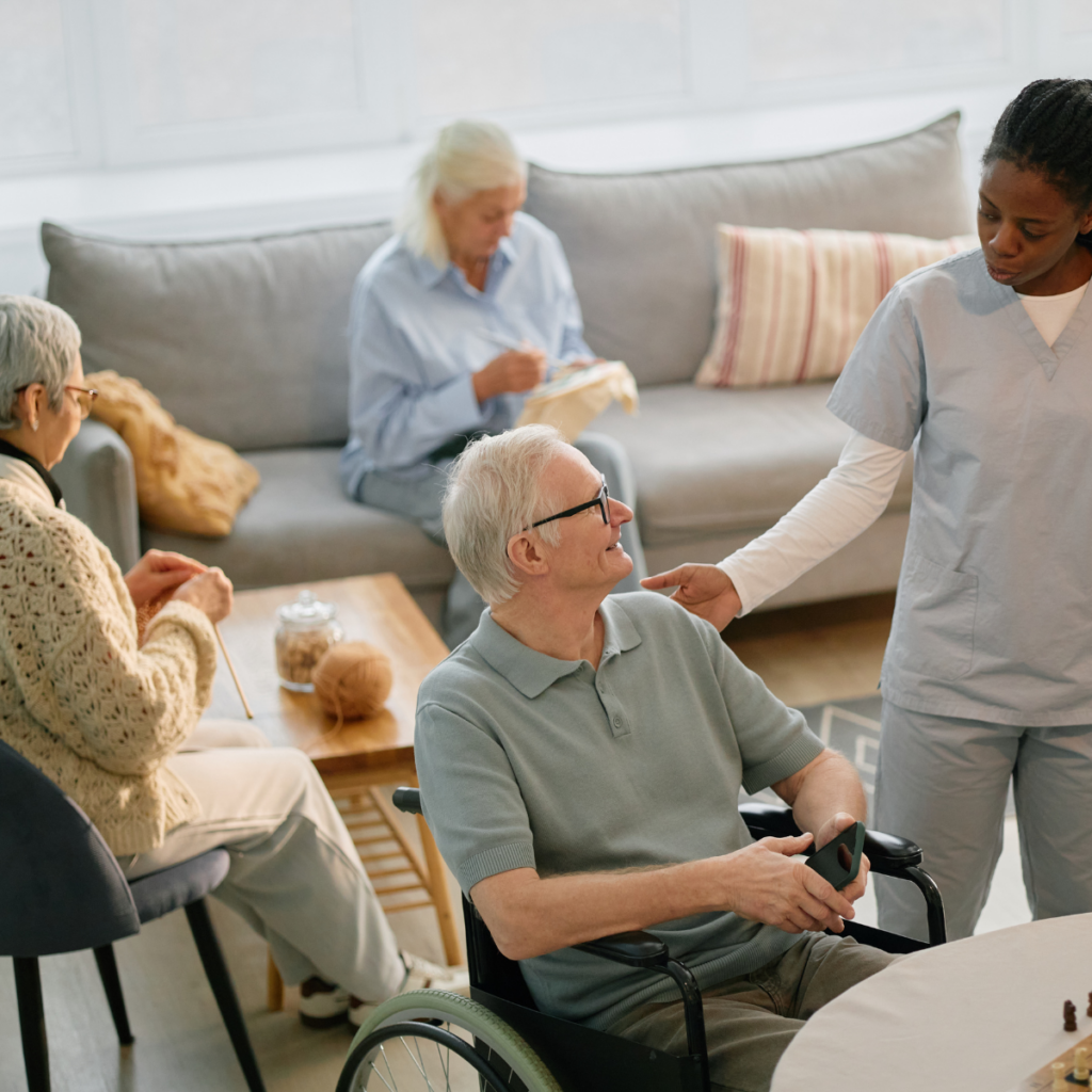 A caregiver in scrubs warmly interacting with an elderly resident in a wheelchair, while other seniors relax and knit in a comfortable, homelike aged care lounge.